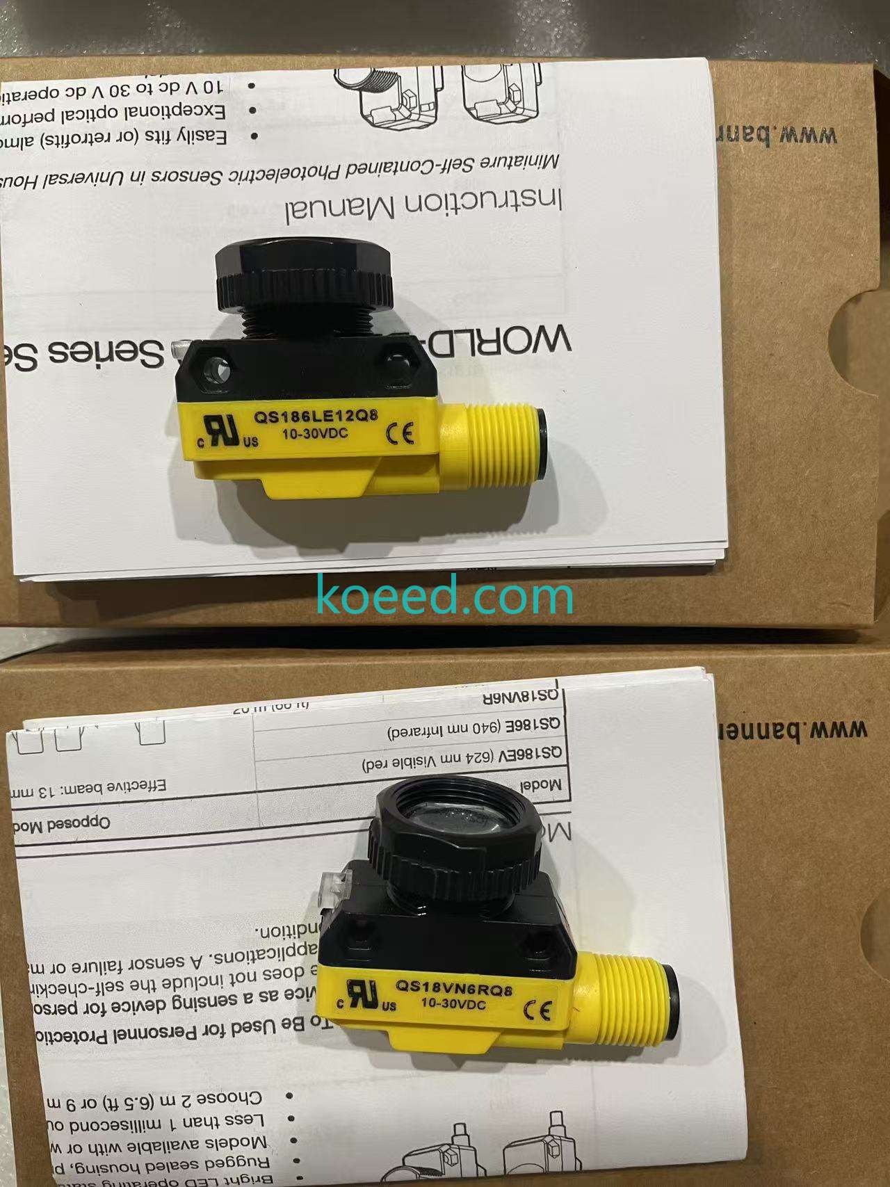 Two yellow-and-black devices marked QS186LE12Q8 and QS18VN6RQ8, each with 10-30VDC wording, resting on instruction sheets and cartons; original manufacture is not authenticated.