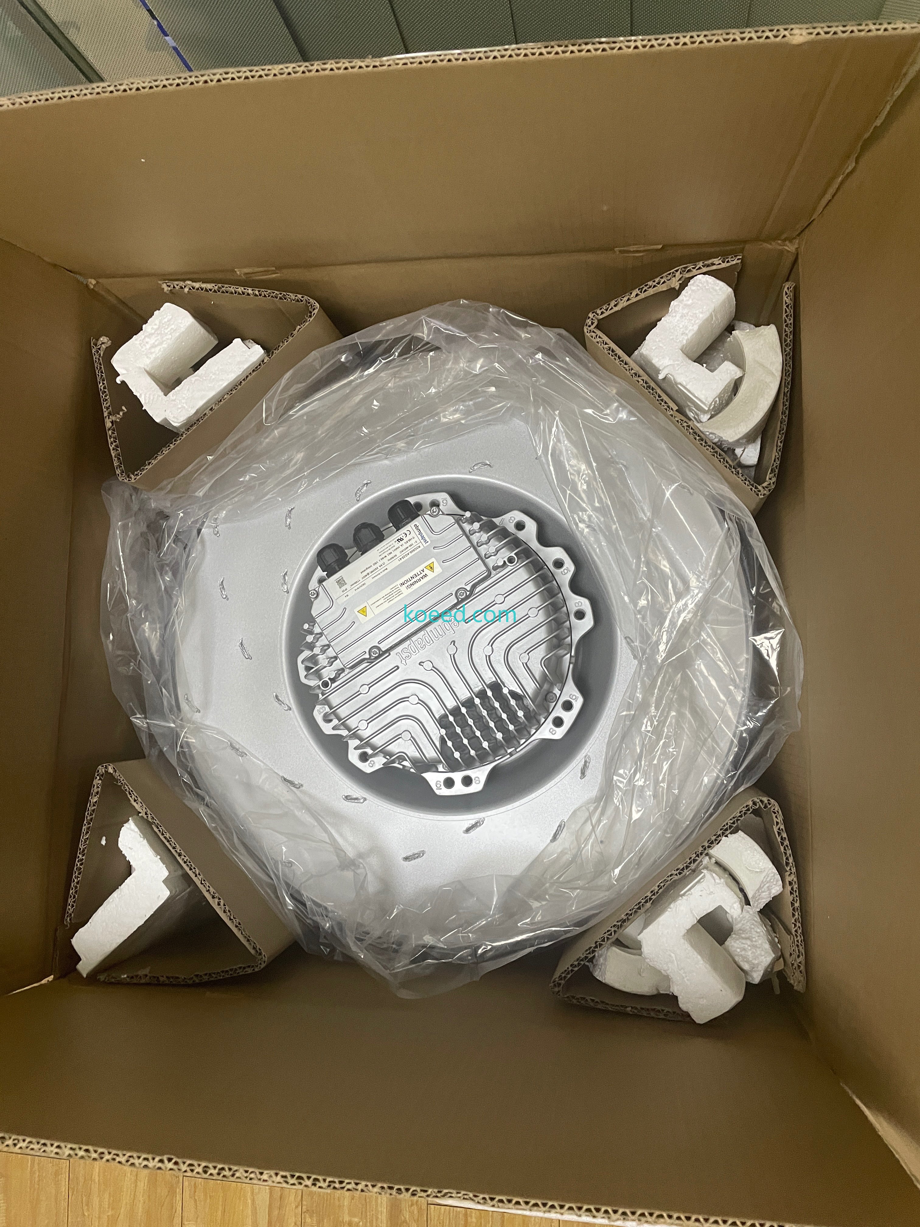 Silver fan assembly in a clear bag inside a carton, with foam and folded-cardboard corner supports, a finned housing and capped cable entries.